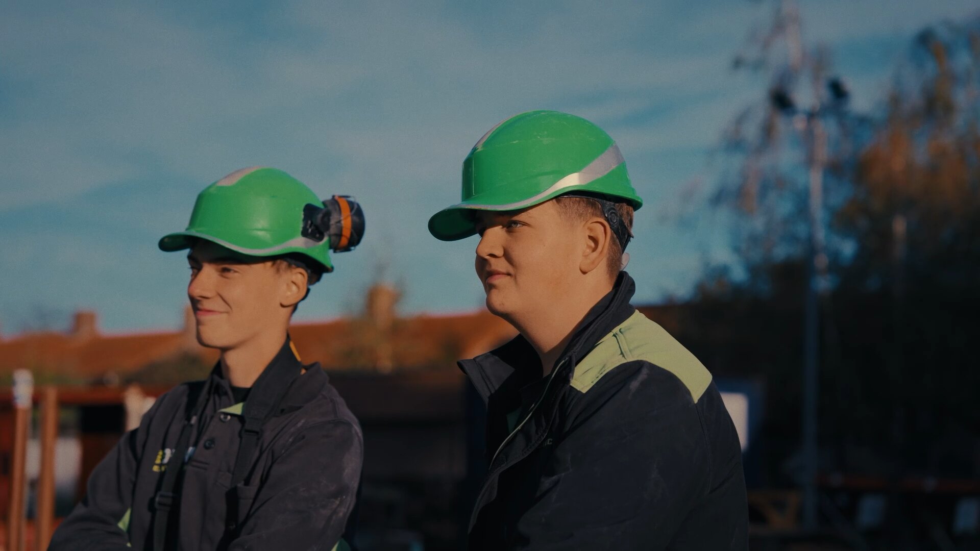 Twee jonge monteurs met groene bouwhelmen kijken lachend uit over de bouwplaats.
