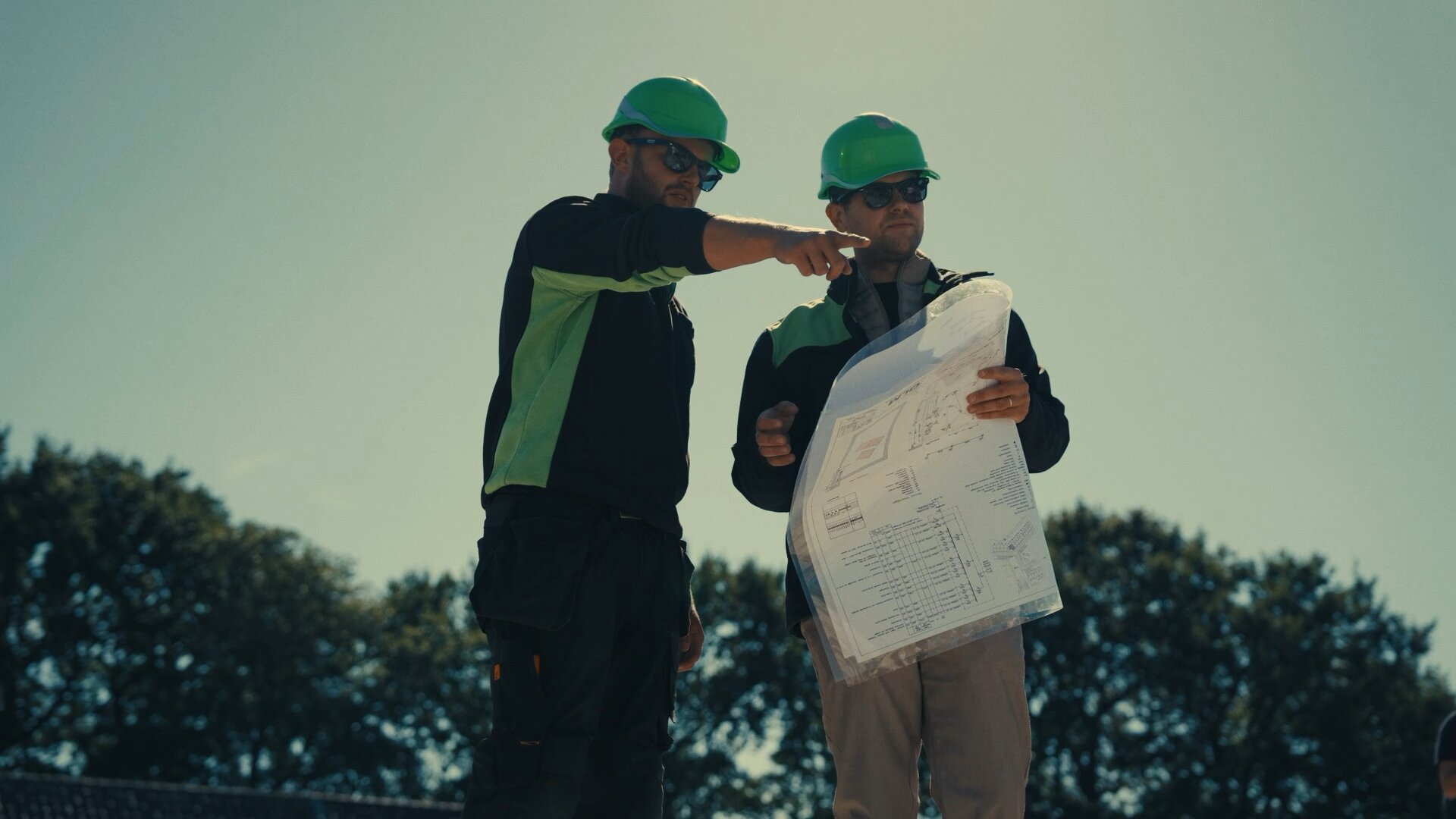 Twee monteurs met groene helmen bekijken een installatietekening, een van hen wijst iets aan.
