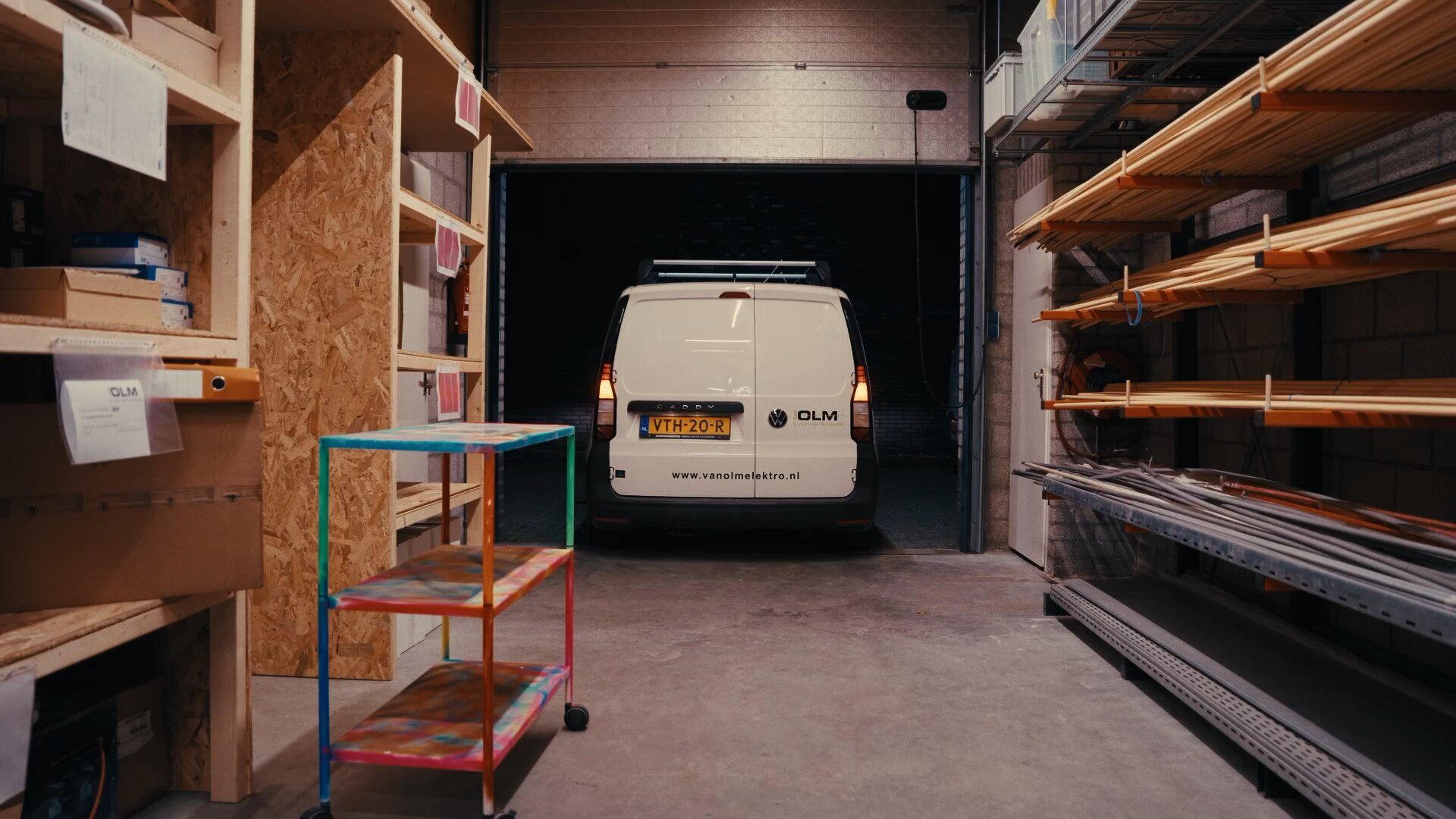 Witte bus van Van Olm met www.vanolmelektro.nl in de werkplaats tussen materiaalrekken.