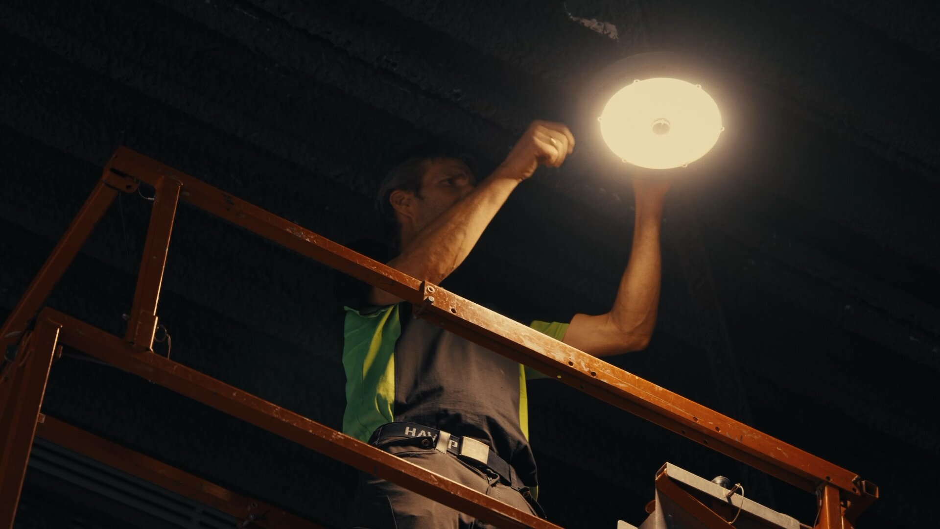 Monteur op een hoogwerker monteert een ronde lamp tegen een donker plafond.