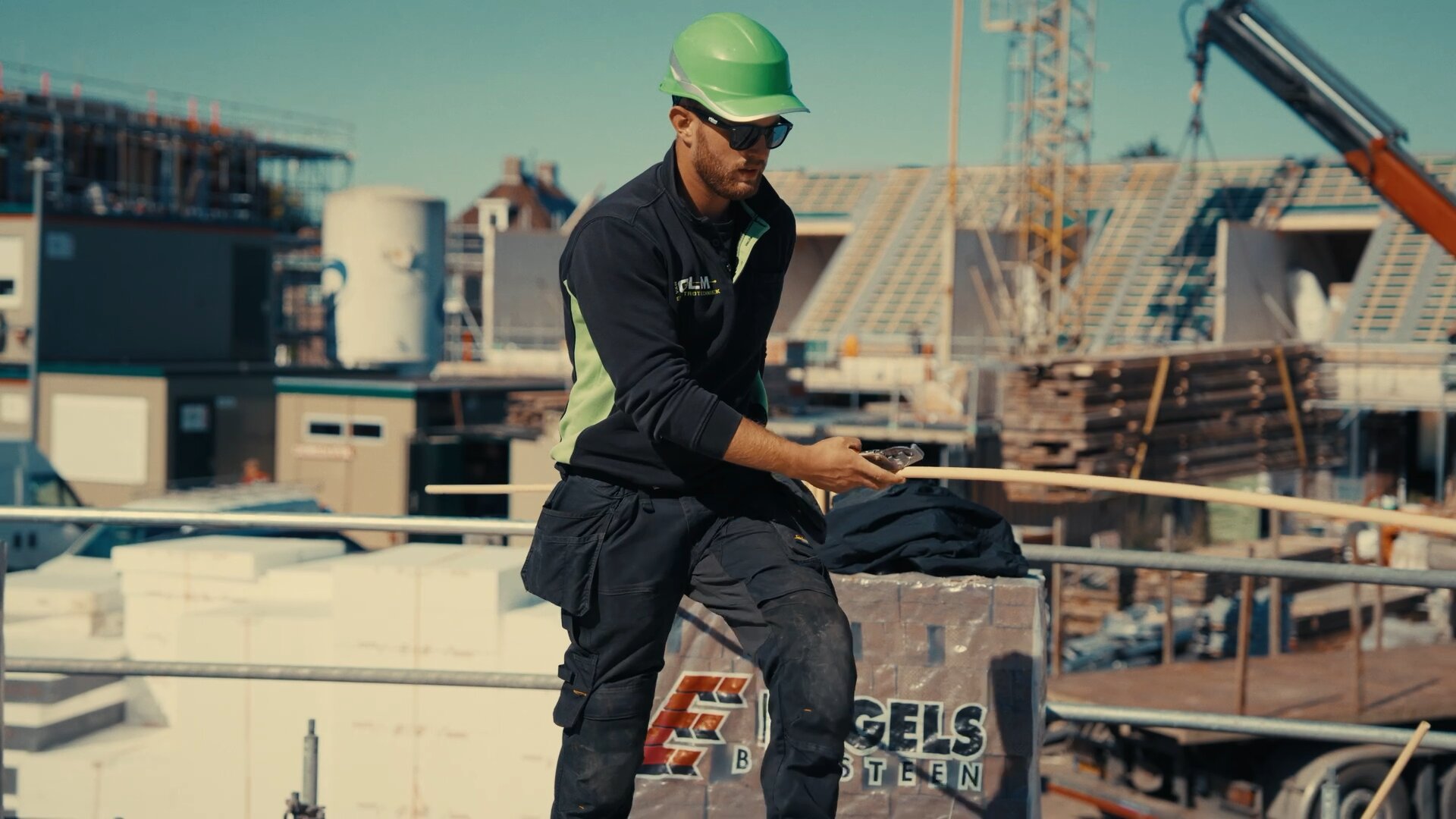 Monteur van Van Olm met groene helm en installatiebuis op een bouwplaats met nieuwbouwwoningen.