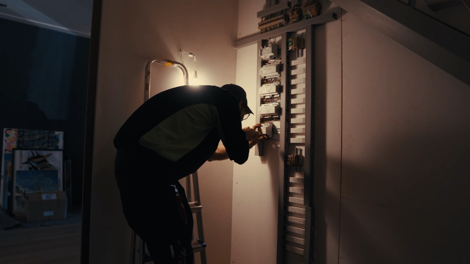 Monteur op een trapje bedraadt modules op een lange installatierail, bijgelicht door een bouwlamp.