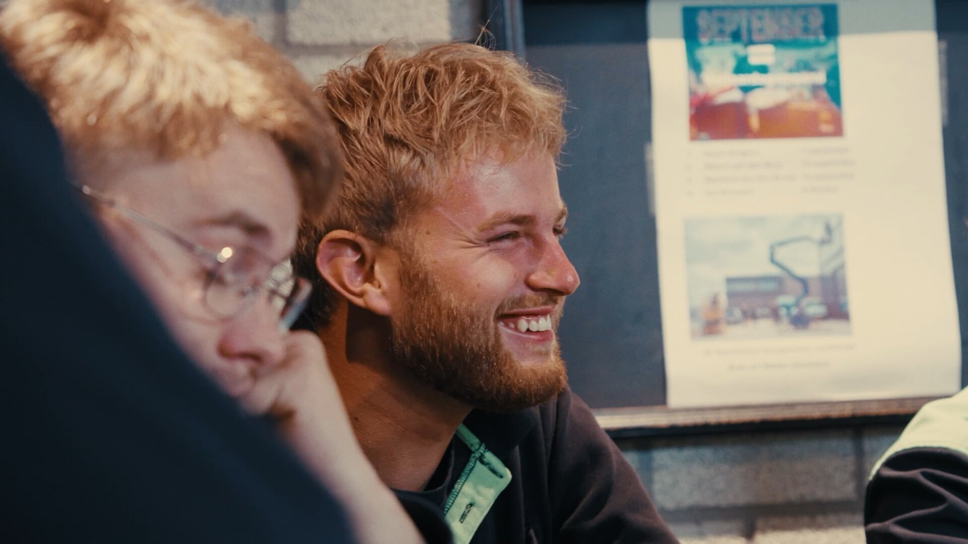 Lachende collega met baard zit naast een collega met bril bij het prikbord.